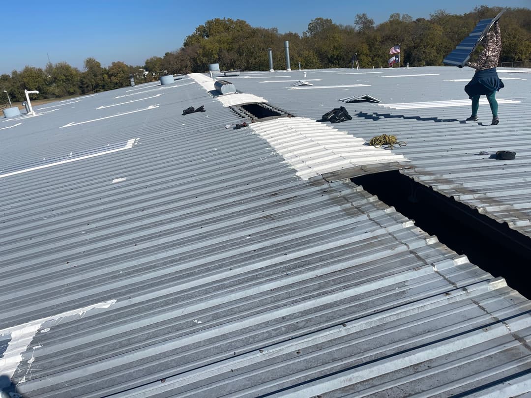 Worker repairing a metal roof with sections removed, clear sky in the background.