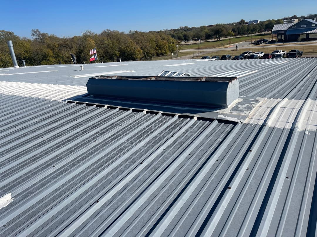 Metal roof with skylight and clear blue sky, surrounded by trees and vehicles below.